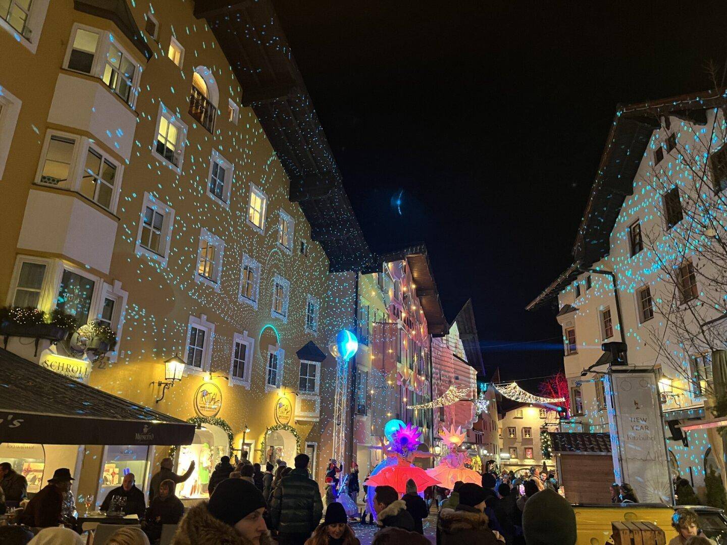 Festlich beleuchtete Gasse in einer Stadt bei Nacht mit bunten Lichtprojektionen und Menschen.