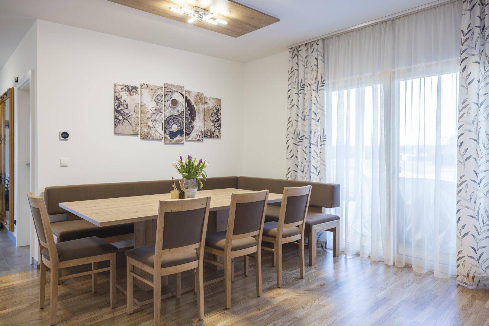 Heller Essbereich mit großem Holztisch und Eckbank, direkt neben einem Fenster mit Vorhängen.