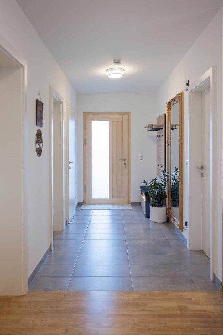 Heller Flur mit modernen grauen Fliesen, einer Holztür und einem großen Wandspiegel.