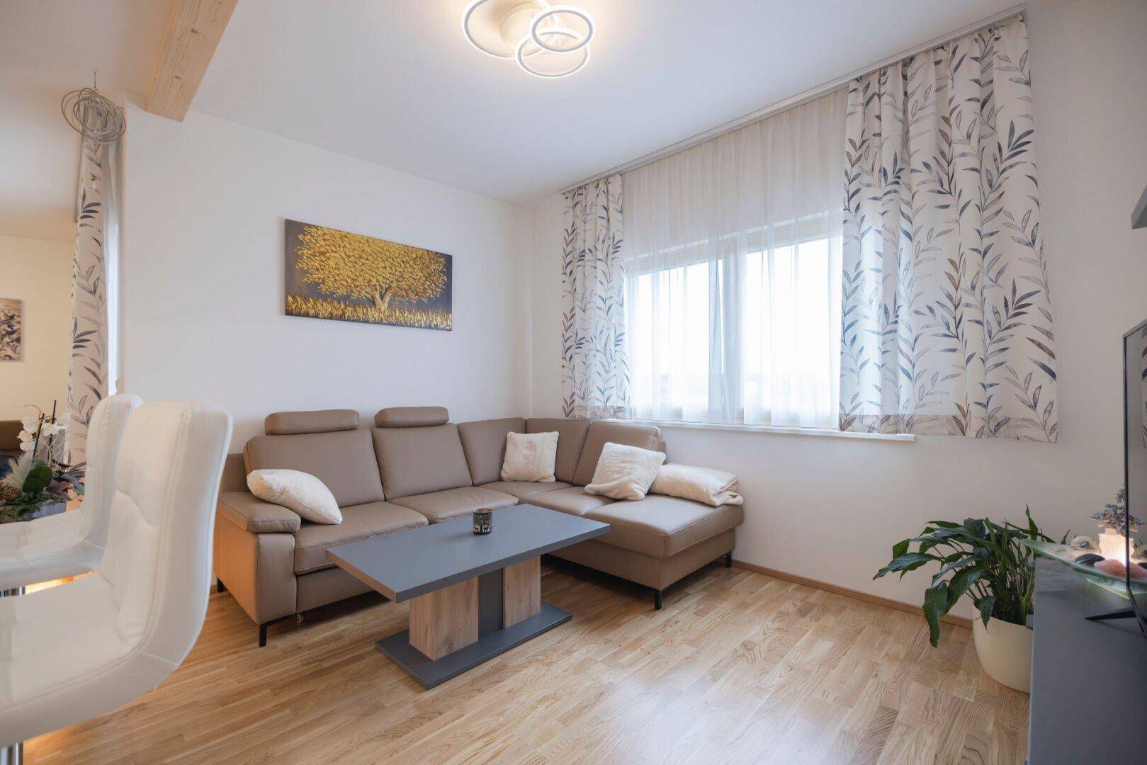 Gemütliches Wohnzimmer mit beigem Ecksofa, Couchtisch und TV-Möbel, gut beleuchtet durch Fenster.