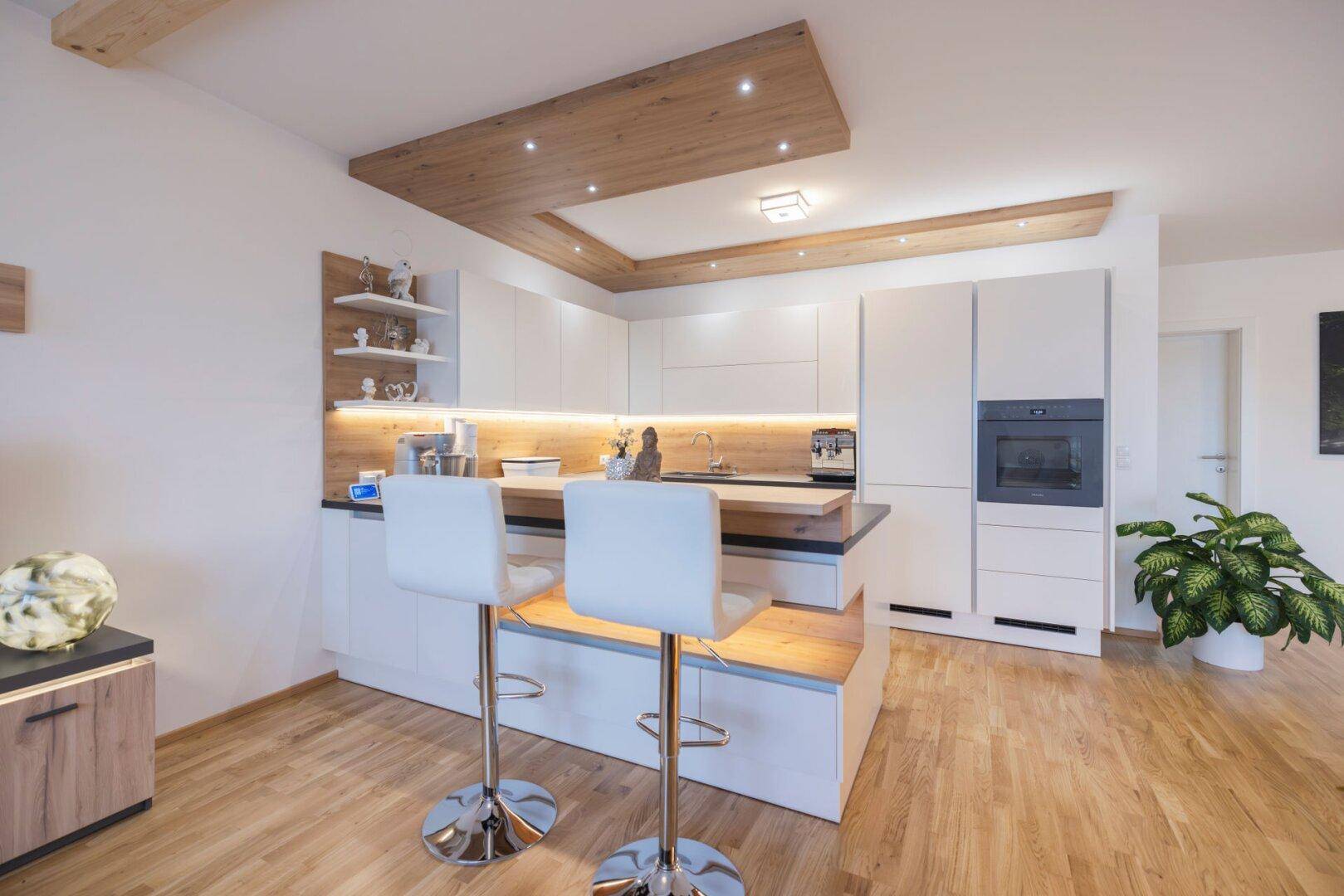 Moderne Einbauküche mit weißen Fronten, Kücheninsel und integrierter Frühstücksbar unter Holzbalken.