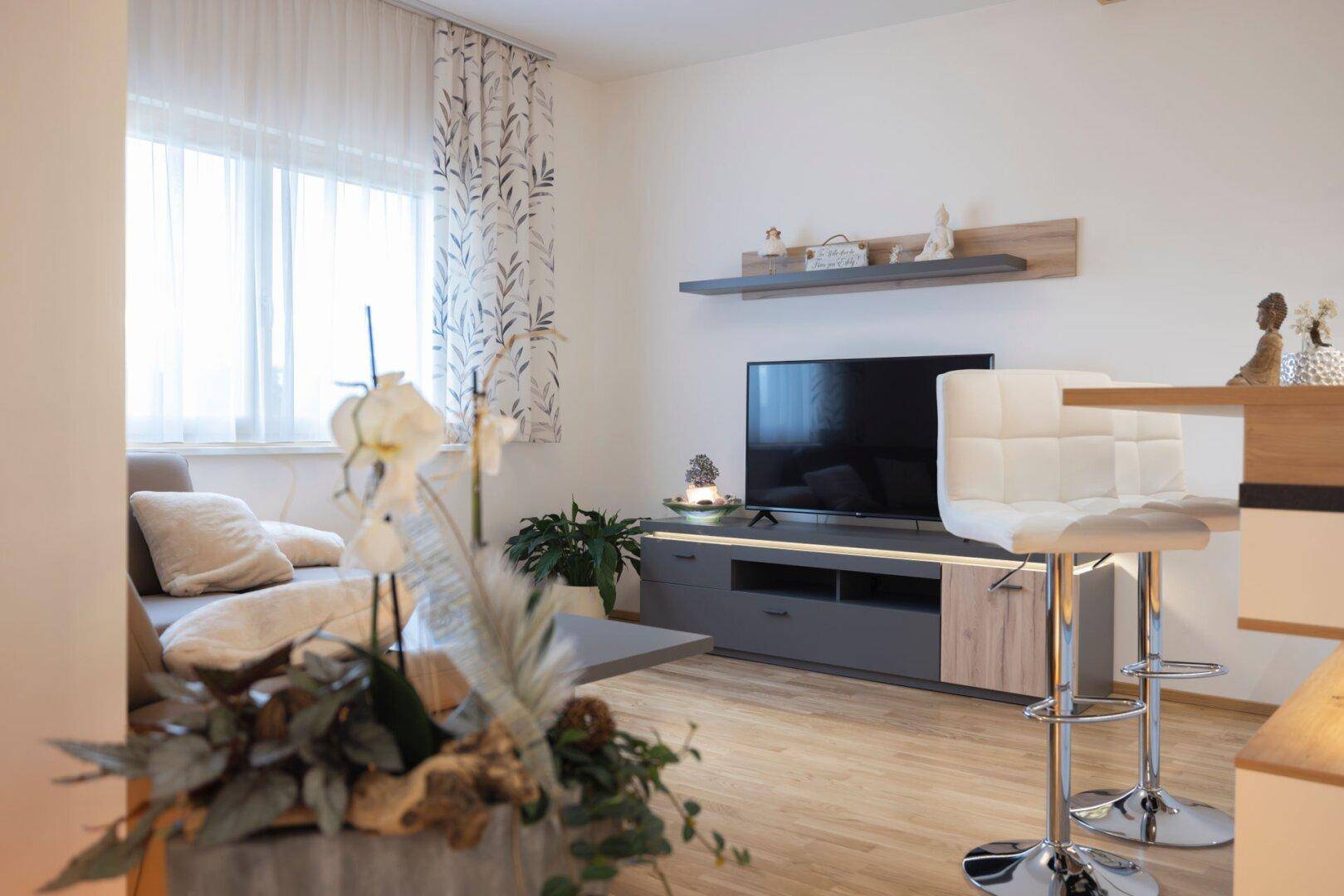 Wohnbereich mit Fernseher, Medienmöbeln und Blick auf die angrenzende Küche mit Frühstücksbar.