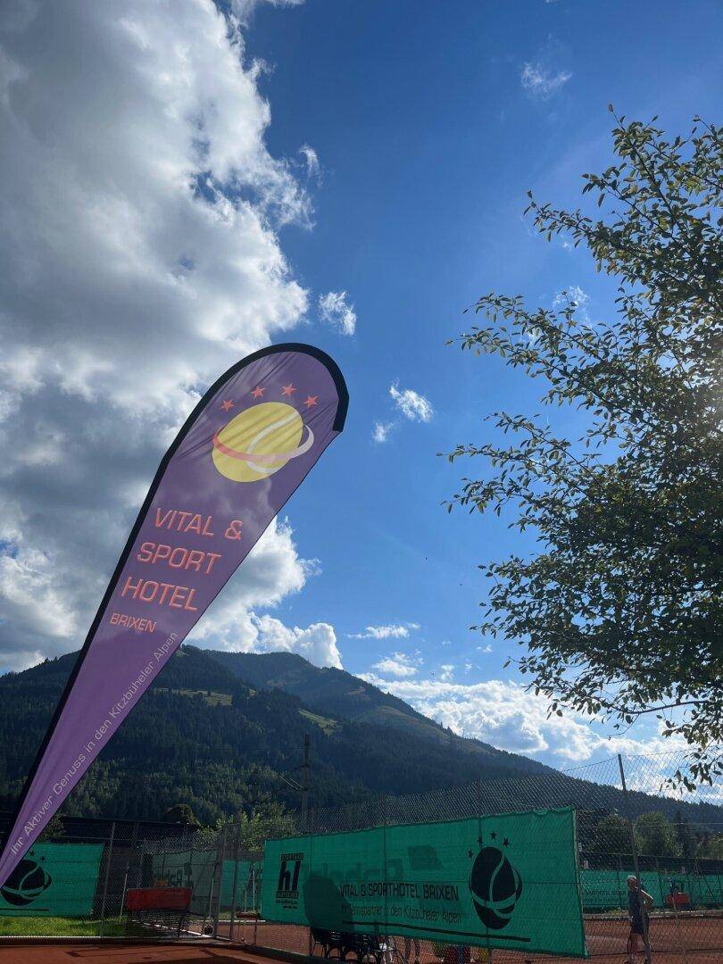Tennisplatz im Freien mit Bergblick und Werbung für ein Vital- und Sporthotel.
