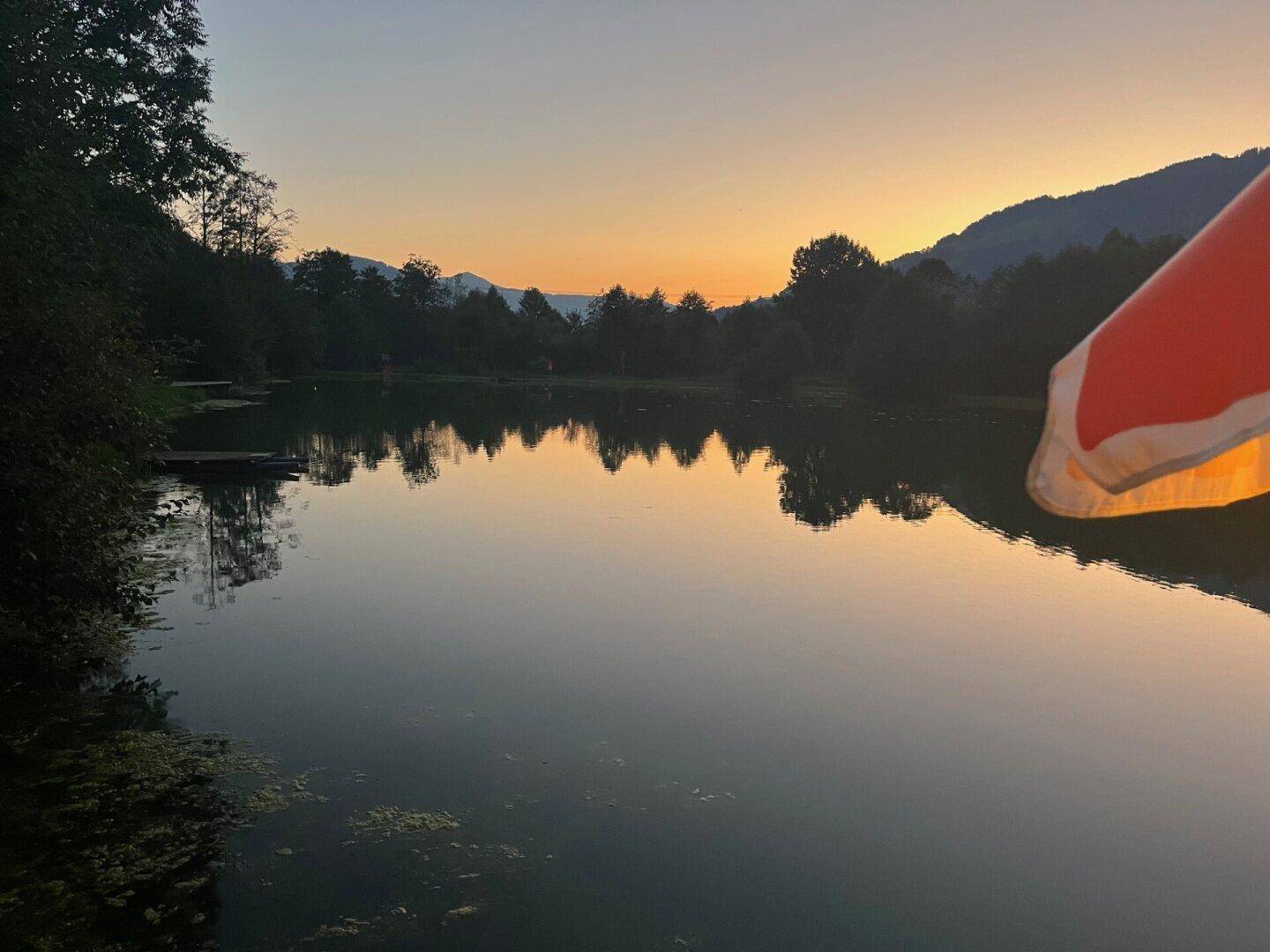 Ruhiger See bei Sonnenuntergang mit Spiegelungen der Bäume und Berge am Horizont.