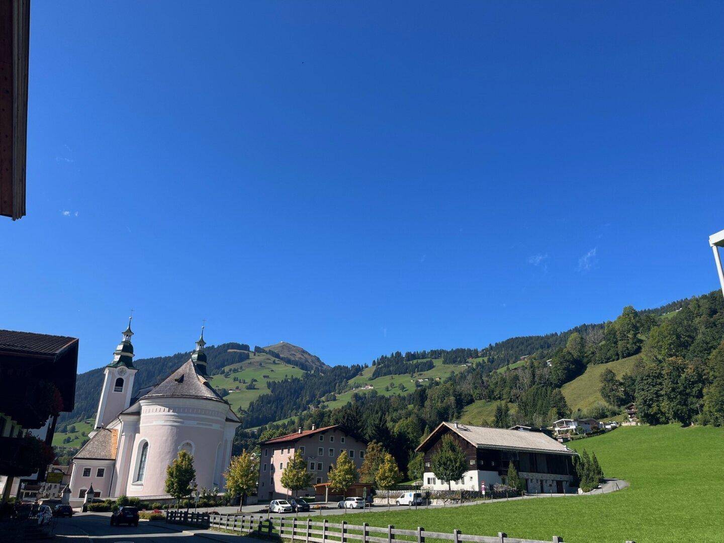 Idyllische Dorfansicht mit Kirche, traditionellen Häusern und grünen Wiesen vor Bergkulisse.
