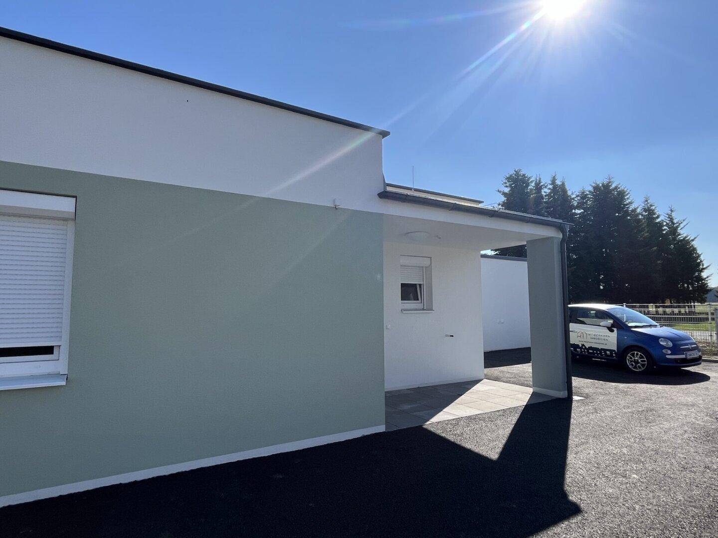 Seitenansicht eines modernen Neubaus mit grüner Fassade und einem überdachten Carport, geparkt ist ein blaues Auto.