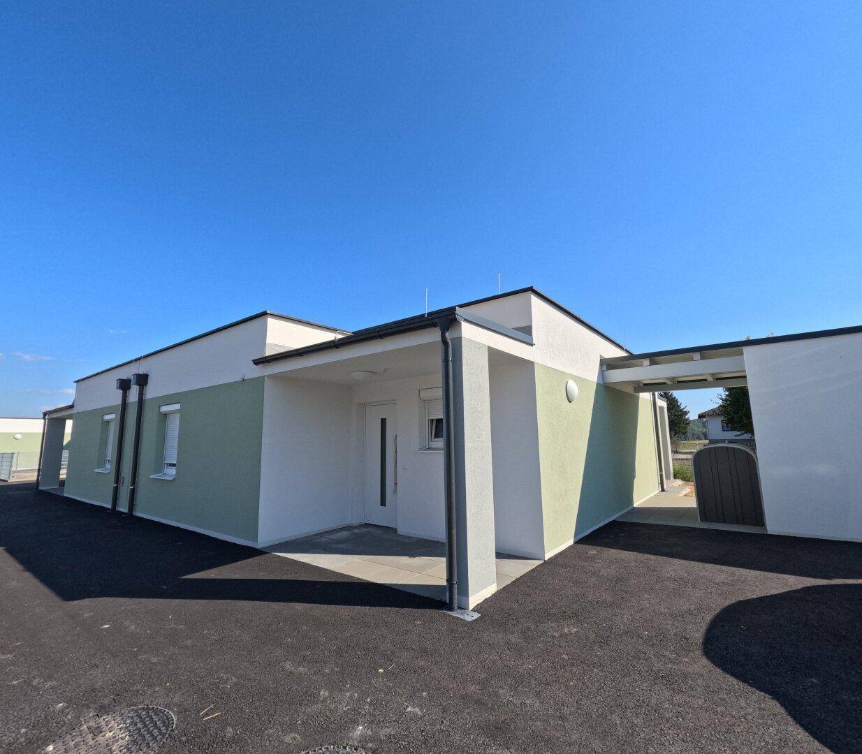 Moderner Neubau mit weißer und grüner Fassade, Eingangsbereich und überdachtem Carport unter strahlendem Himmel.