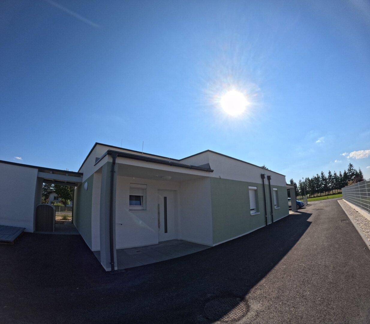 Moderner Neubau mit weißer und grüner Fassade, Eingangsbereich und überdachtem Carport unter strahlendem Himmel.