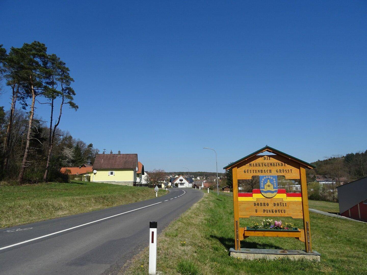 Landstraße führt in eine ländliche Gemeinde mit Willkommensschild und grüner Umgebung.