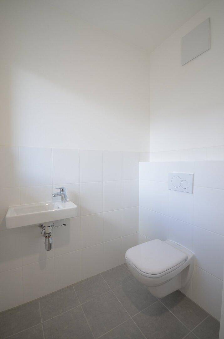 Helles Gäste-WC mit Wand-WC, kleinem Waschbecken und grauen Bodenfliesen.