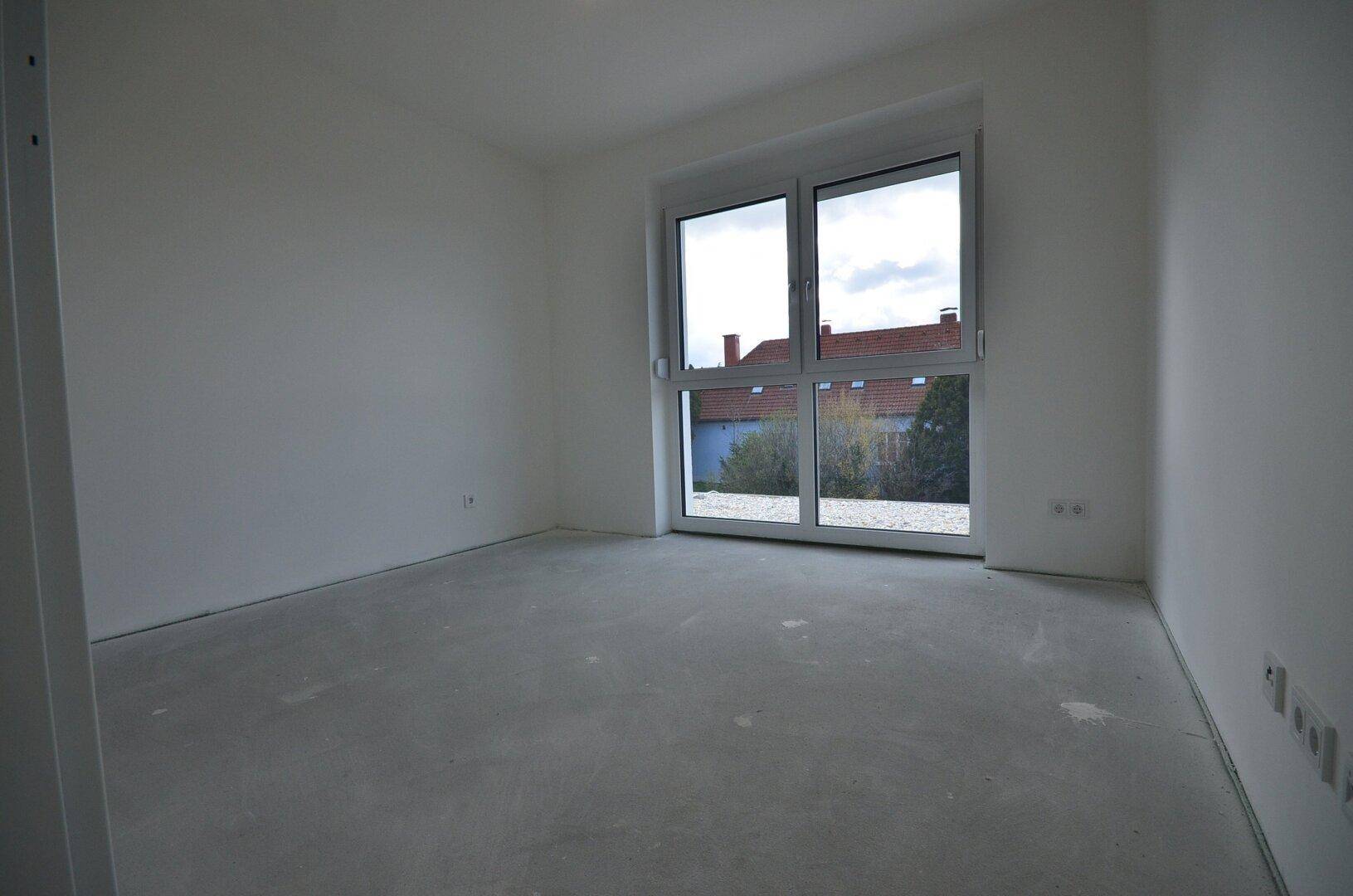 Helles Schlafzimmer im Rohbauzustand mit großem Fenster und Blick auf die Umgebung.