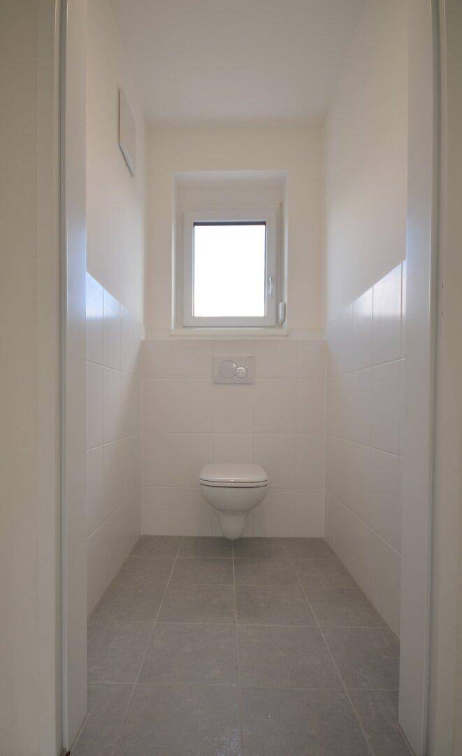 Schlichtes Gäste-WC mit Wand-WC, Fenster und hellgrauen Bodenfliesen.