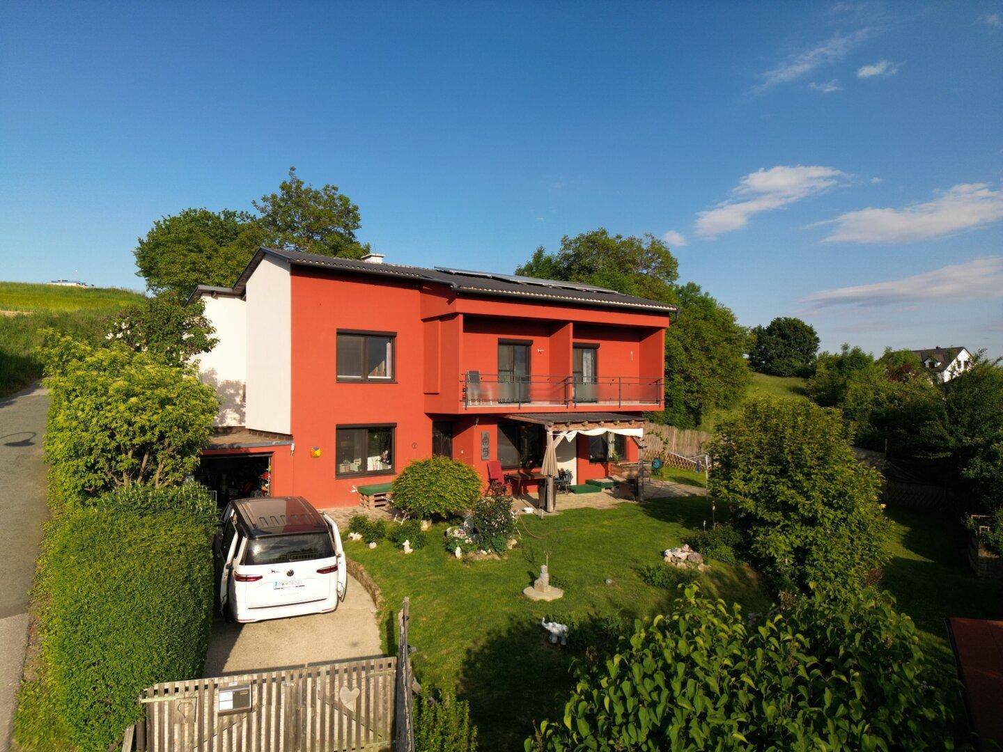 Rotes Wohnhaus mit Balkonen, Solaranlage und Zufahrt in ländlicher Umgebung.