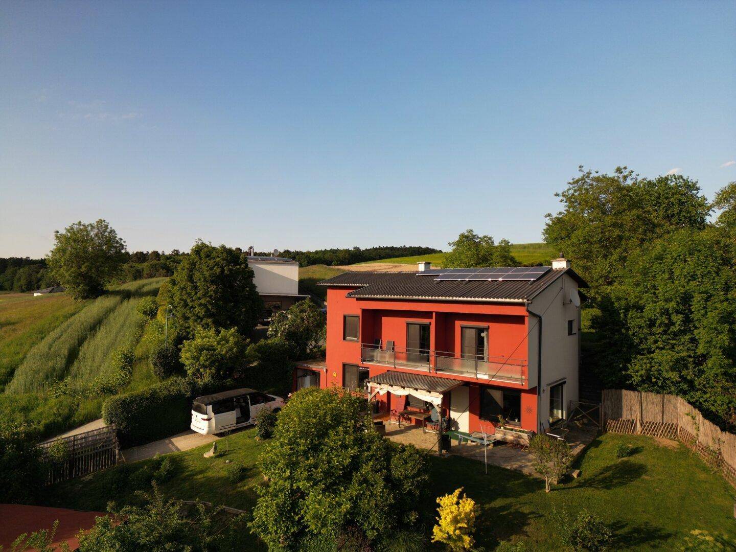 Luftaufnahme eines roten Hauses mit Solaranlage auf dem Dach und umgebender Natur.