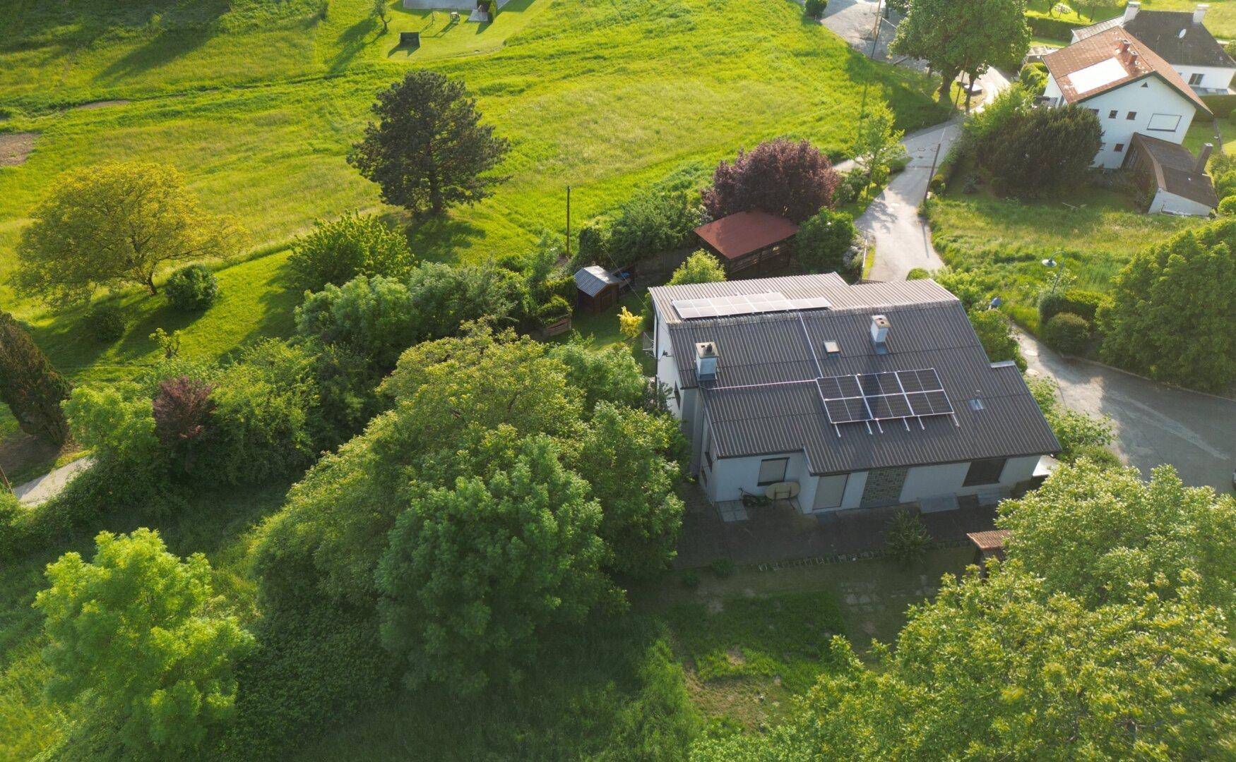 Weitläufige Luftaufnahme eines Hauses mit Solaranlage, umgeben von grünen Feldern und Bäumen.
