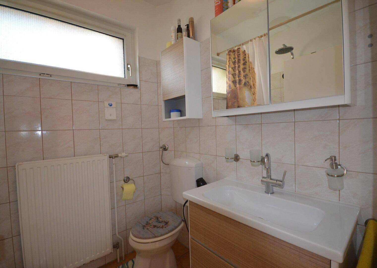 Badezimmer mit Fenster, Waschbecken, Toilette und Spiegelschrank.