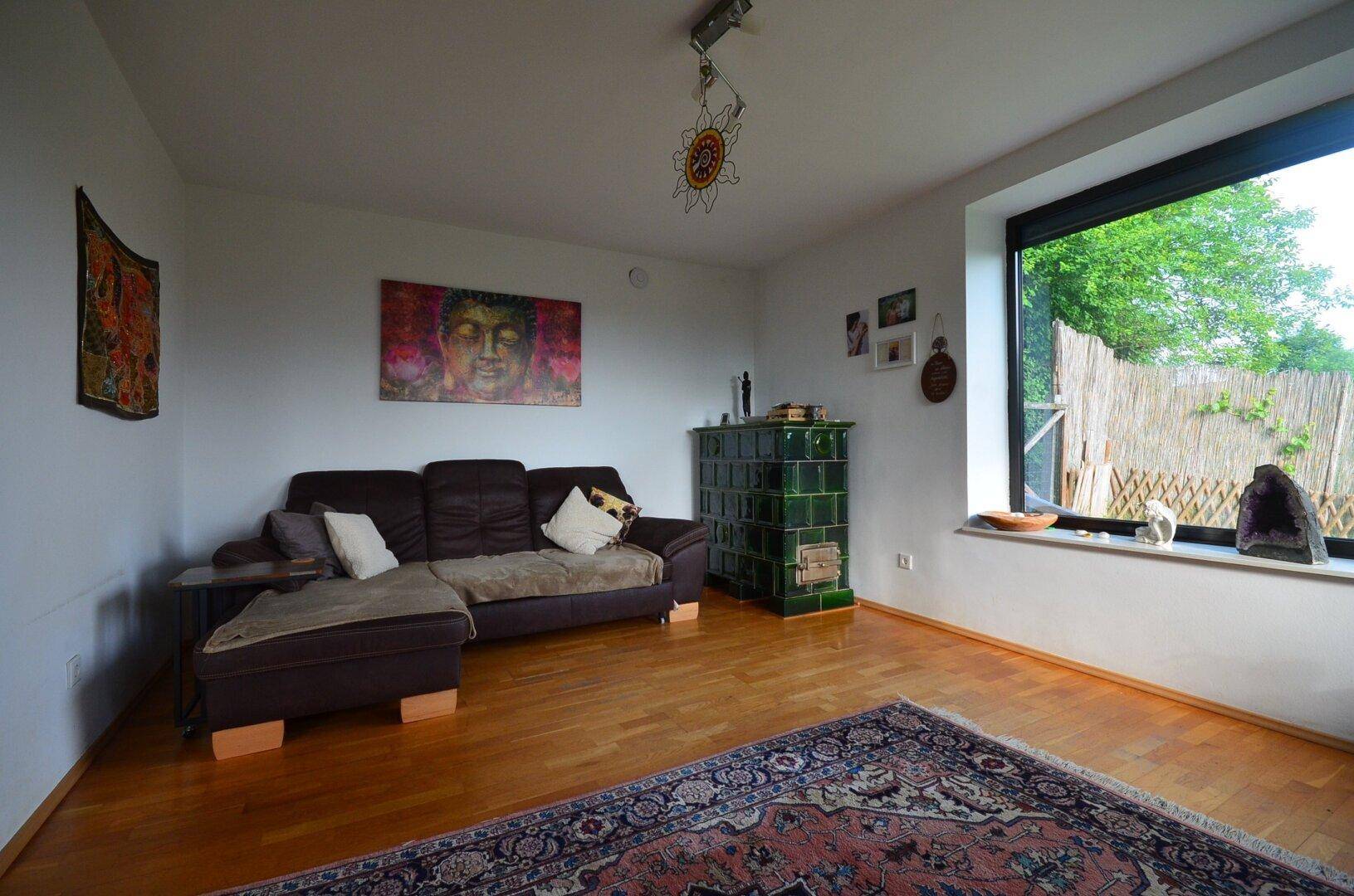 Gemütliches Wohnzimmer mit Kachelofen, Holzboden und großem Fenster zum Garten.