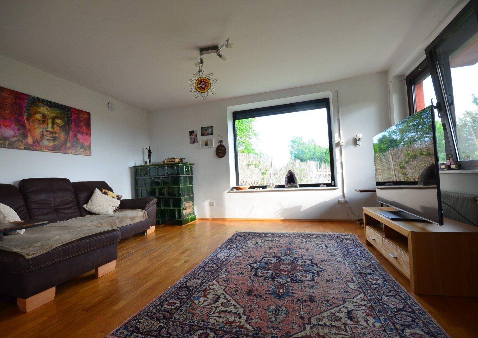 Helles Wohnzimmer mit Holzboden, Kachelofen und Blick ins Grüne durch große Fenster.