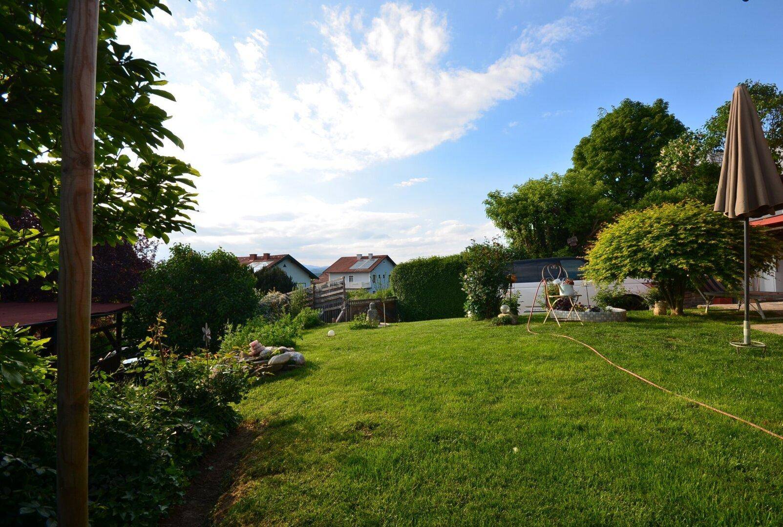 Weitläufiger Garten mit Rasen, Bäumen und einem Sonnenschirm, der eine entspannte Atmosphäre schafft.