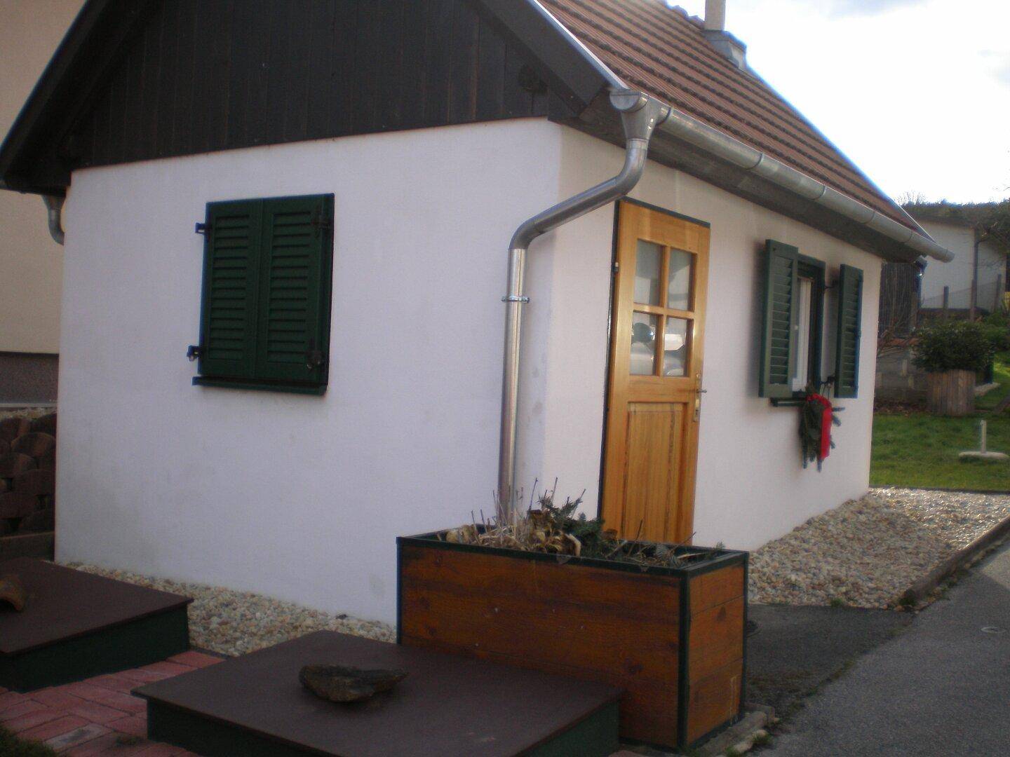 Kleines Nebengebäude mit weißer Fassade, Holztür und grünen Fensterläden im Freien.
