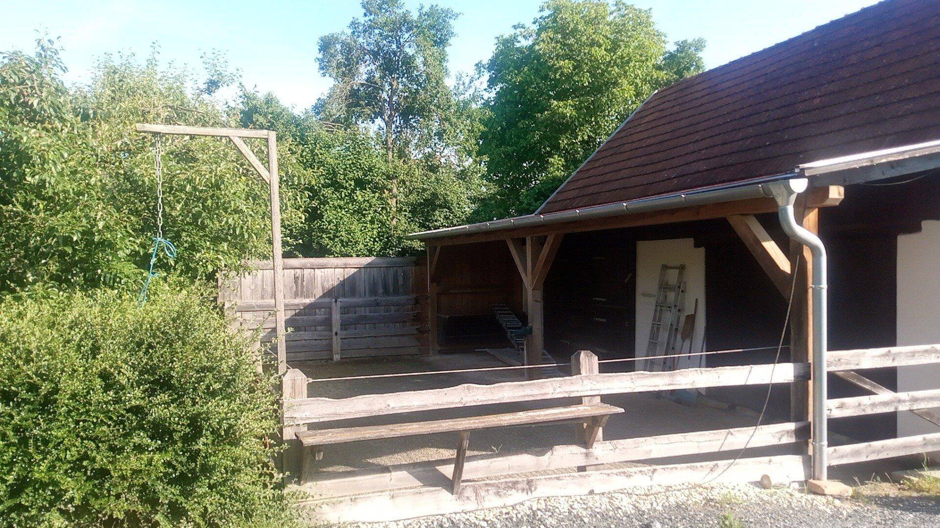 Überdachter Außenbereich mit Holzbänken und einer Schaukel in einem grünen Garten.