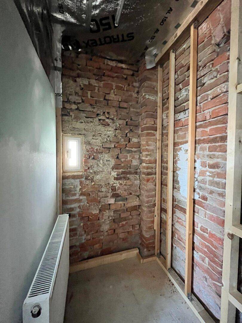 Kleiner Raum im Rohbau mit freiliegenden Ziegelwänden und einem Heizkörper unter dem Fenster.