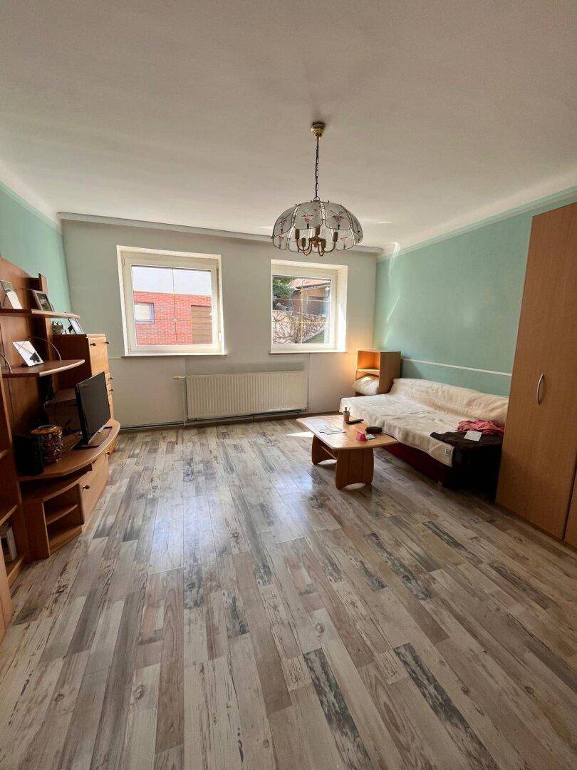 Helles Wohnzimmer mit Laminatboden, Couch und einem Mediamöbel unter zwei Fenstern.