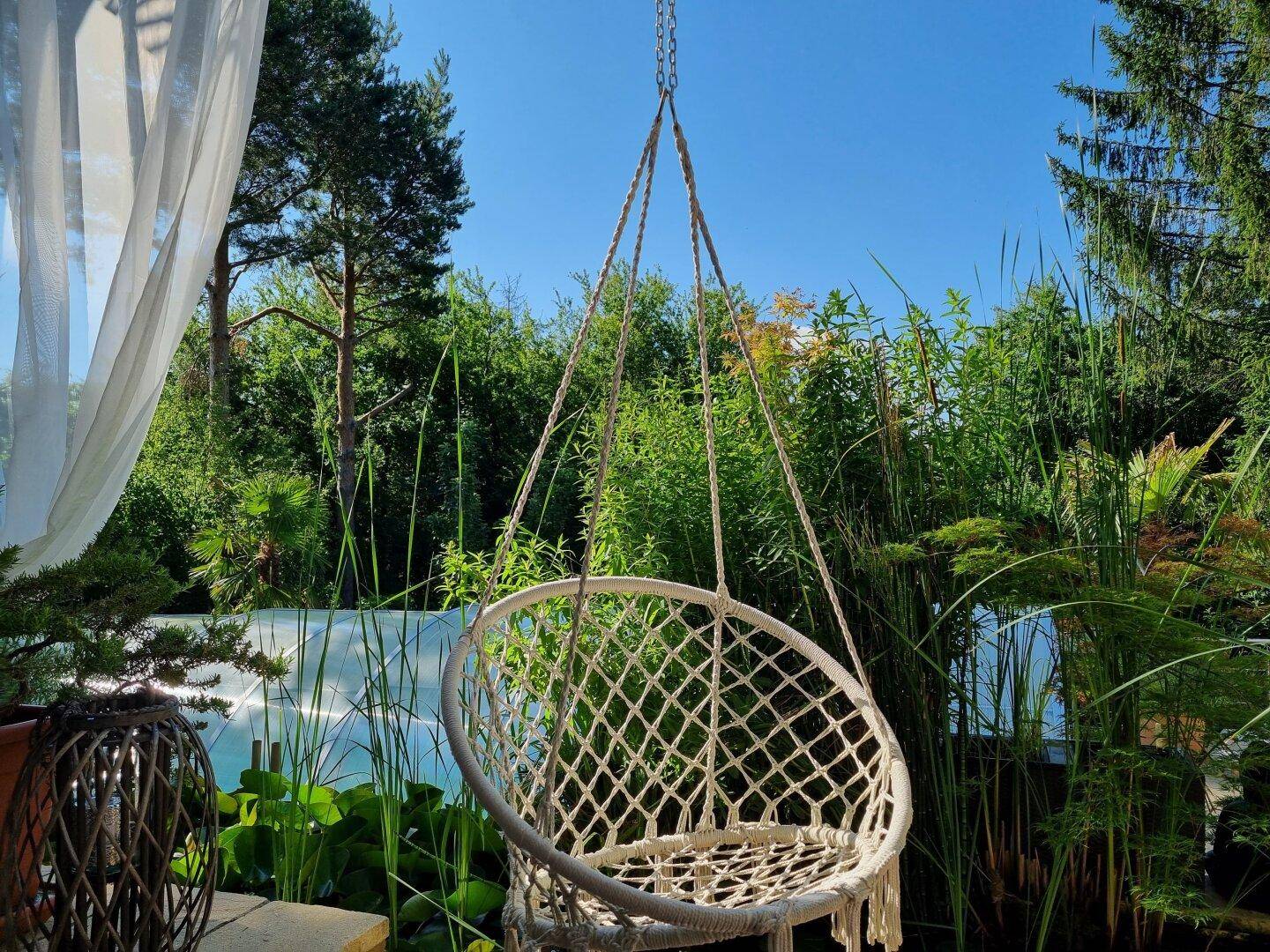 Gemütlicher Hängesessel im Garten, umgeben von dichter grüner Vegetation unter blauem Himmel.