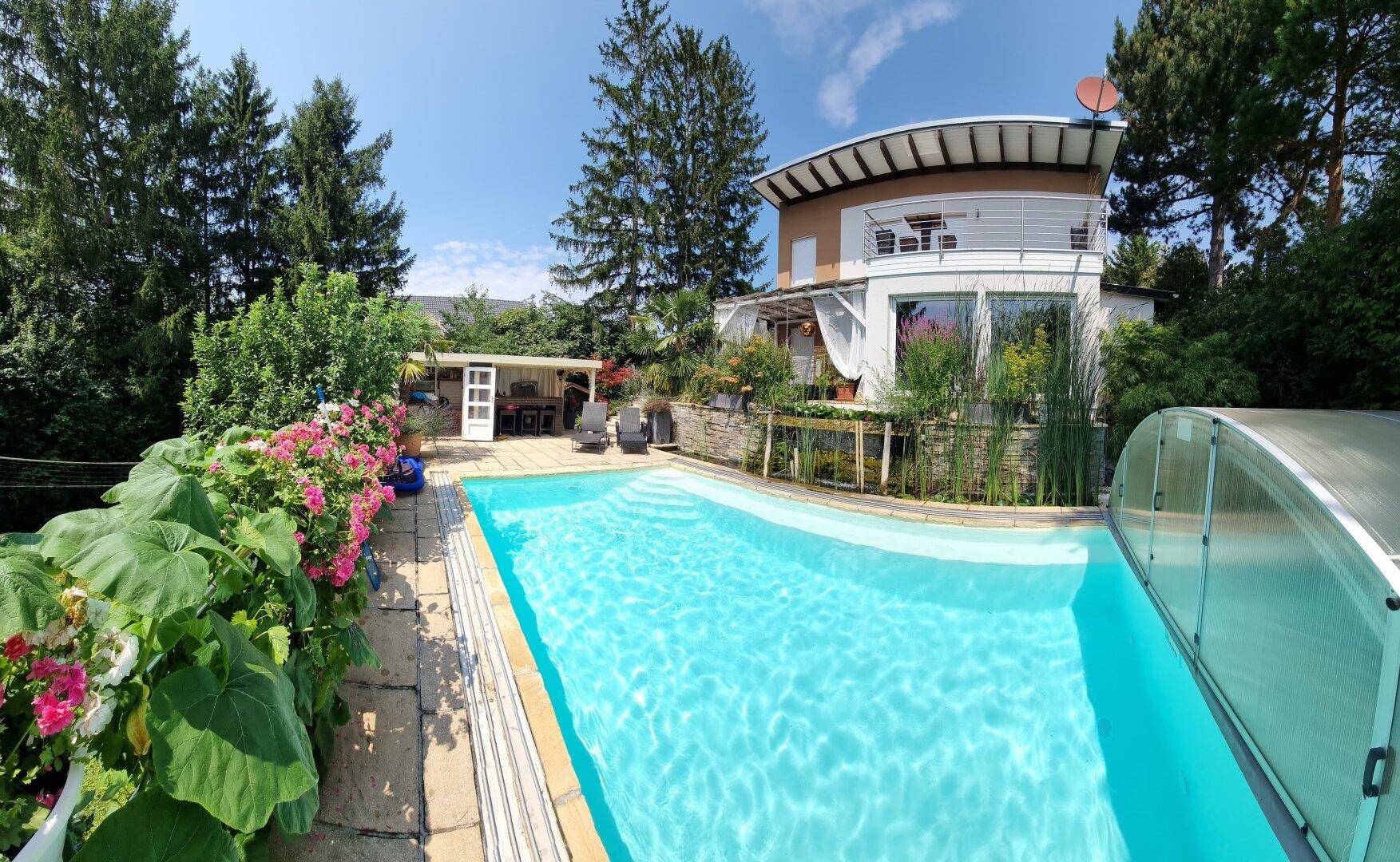 Panoramablick auf das Haus mit großem Pool und blühenden Pflanzen im sonnigen Garten.
