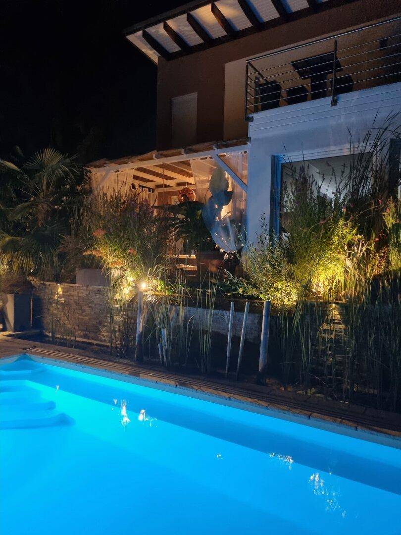 Stimmungsvoll beleuchteter Pool und die Hausfassade bei Nacht, umgeben von hohen Gräsern.