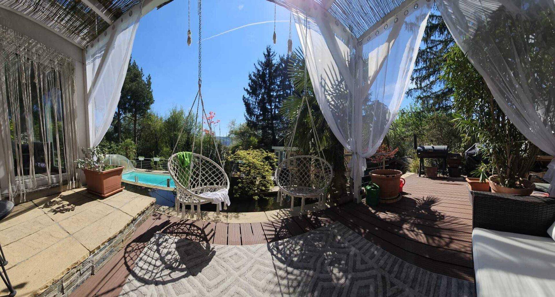 Helle überdachte Terrasse mit Hängesesseln, Teppich und Blick auf den Garten und Pool.