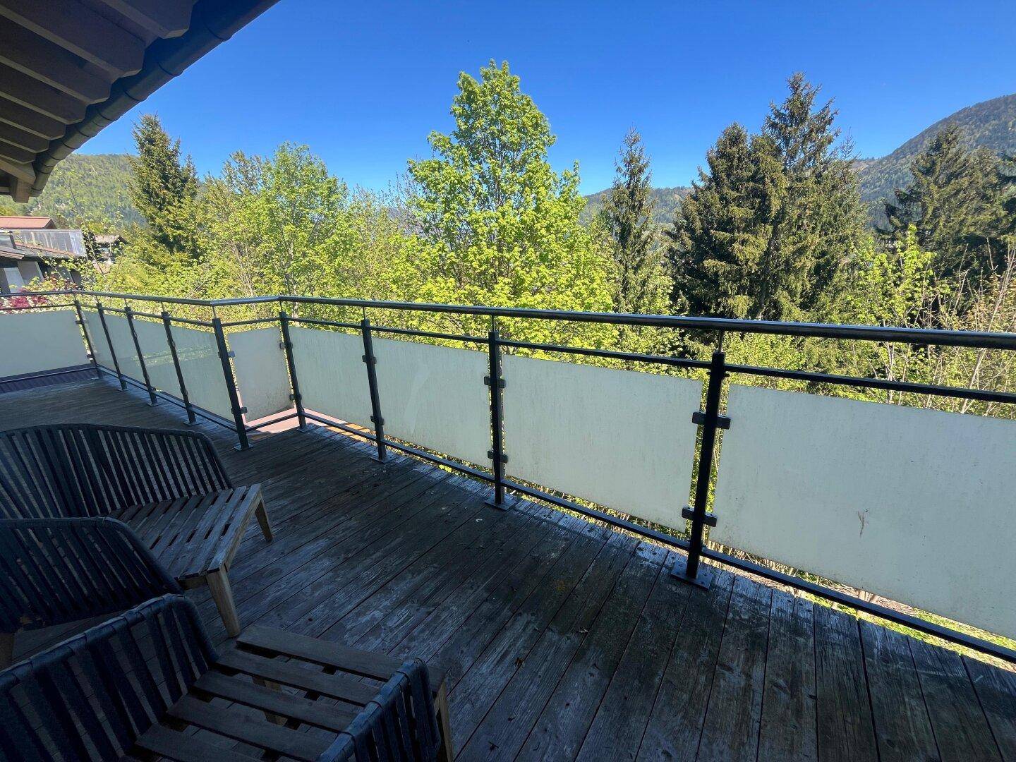 Langer Balkon mit Holzboden und Sitzgelegenheiten, umgeben von grünen Bäumen.