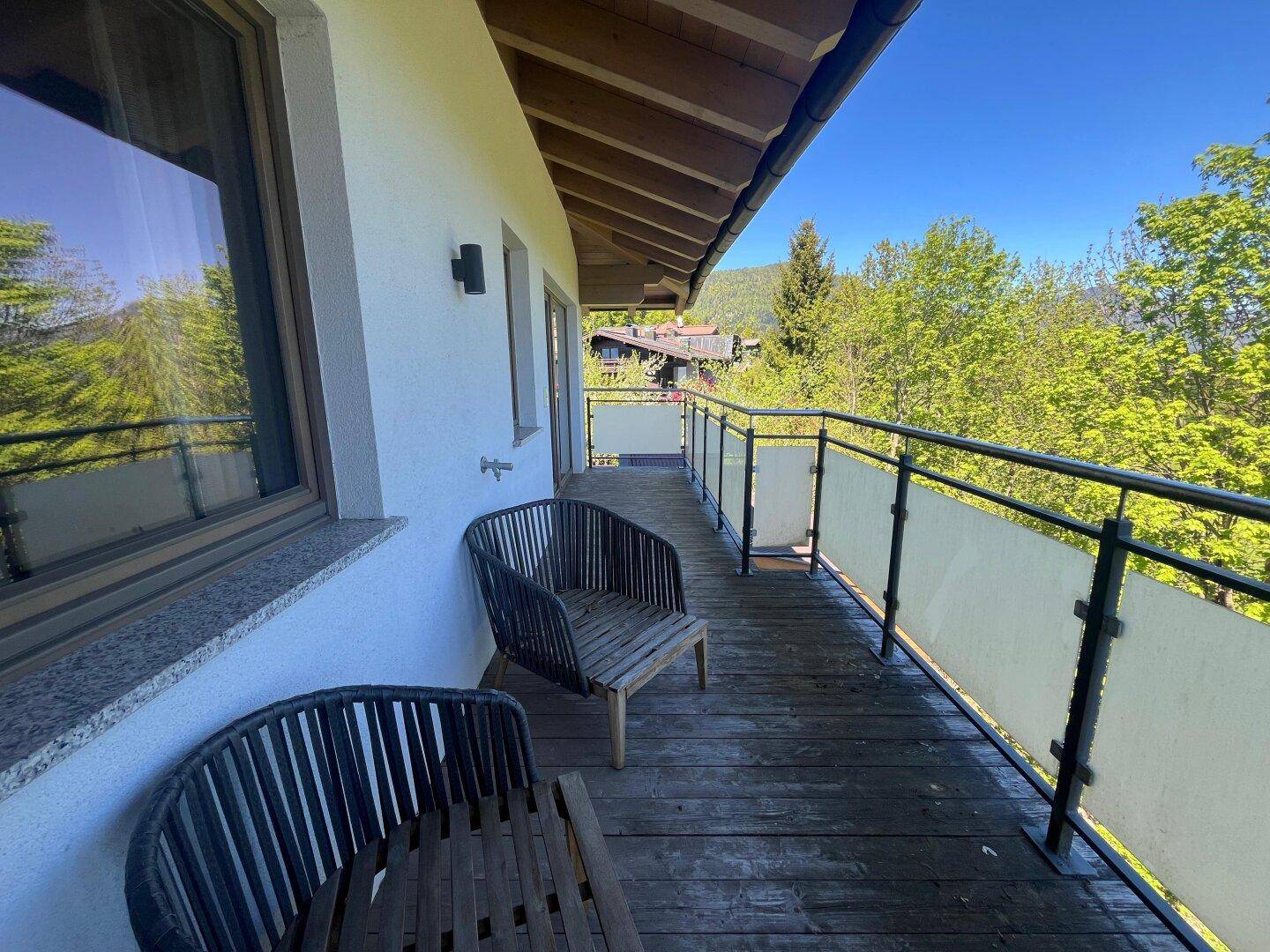 Heller Balkon mit Holzboden und Sitzmöbeln, umgeben von üppiger Vegetation.