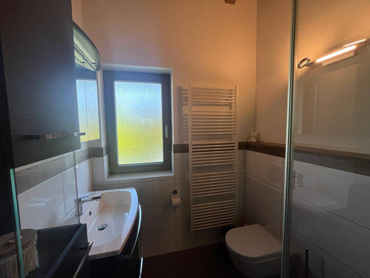 Kleines Badezimmer mit Waschtisch, Toilette und beheiztem Handtuchhalter, hell durch ein Fenster.