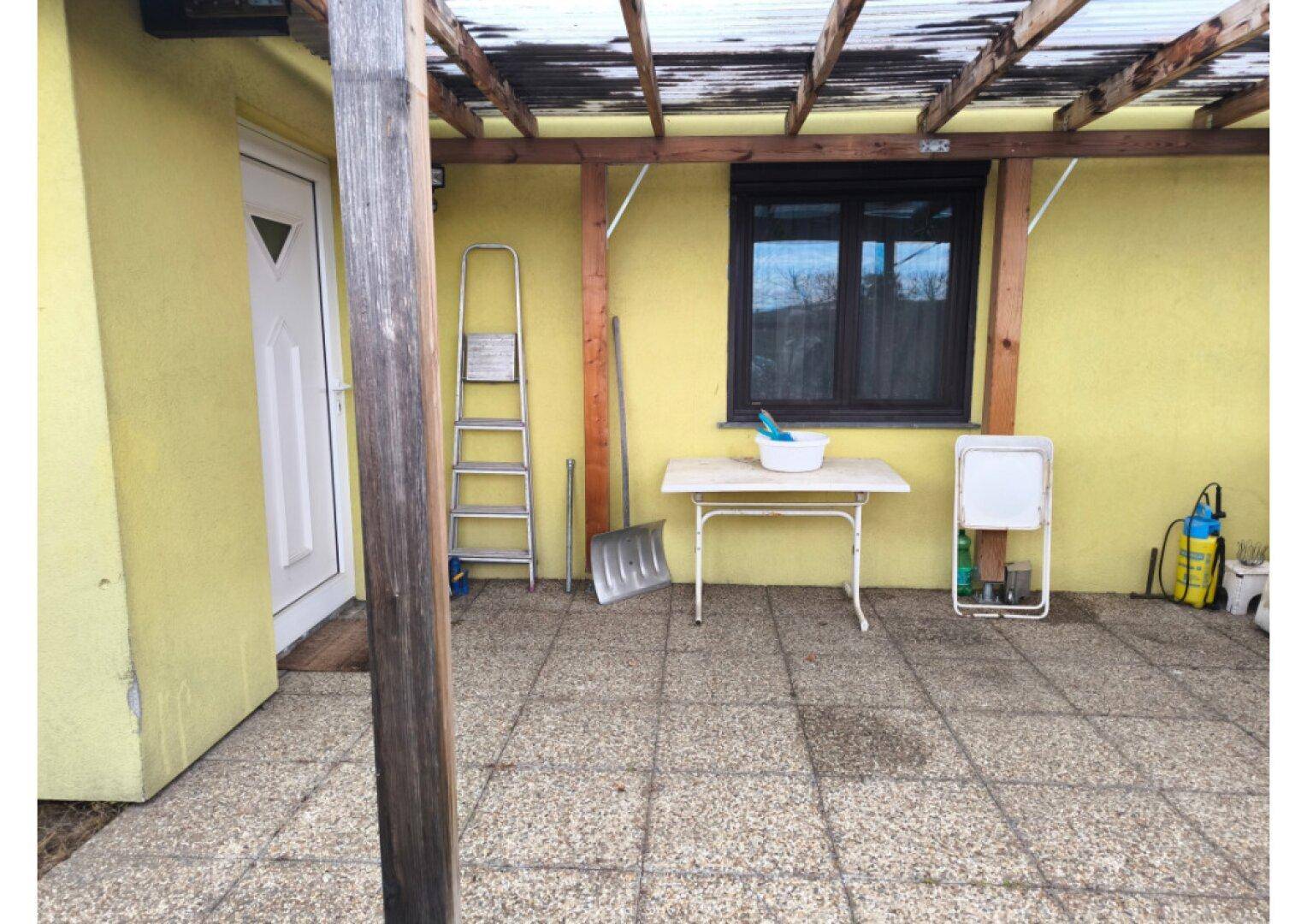 Überdachte Terrasse mit Fliesenboden und gelber Hauswand, die einen praktischen Außenbereich bietet.