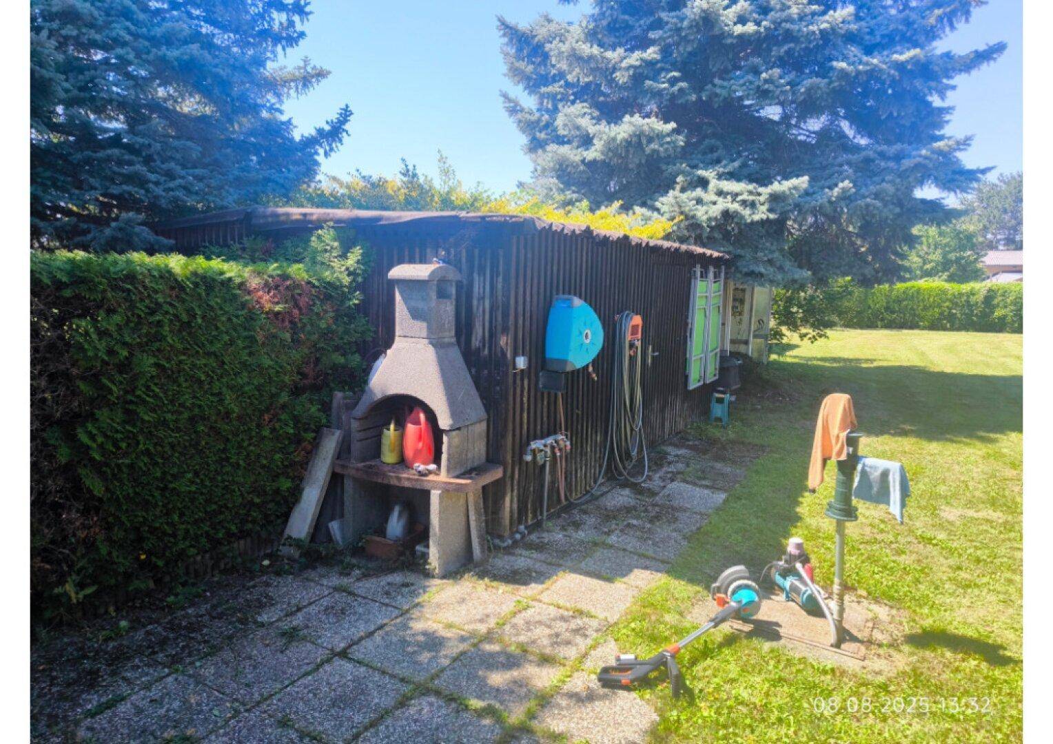Gartenbereich mit gemauertem Grillplatz, Holzschuppen und hohen Bäumen.