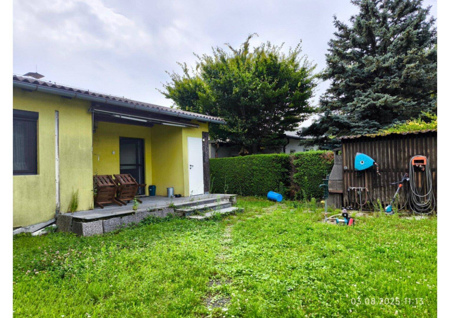 Garten mit Rasenfläche, einer kleinen Terrasse am Haus und einem Holzschuppen.