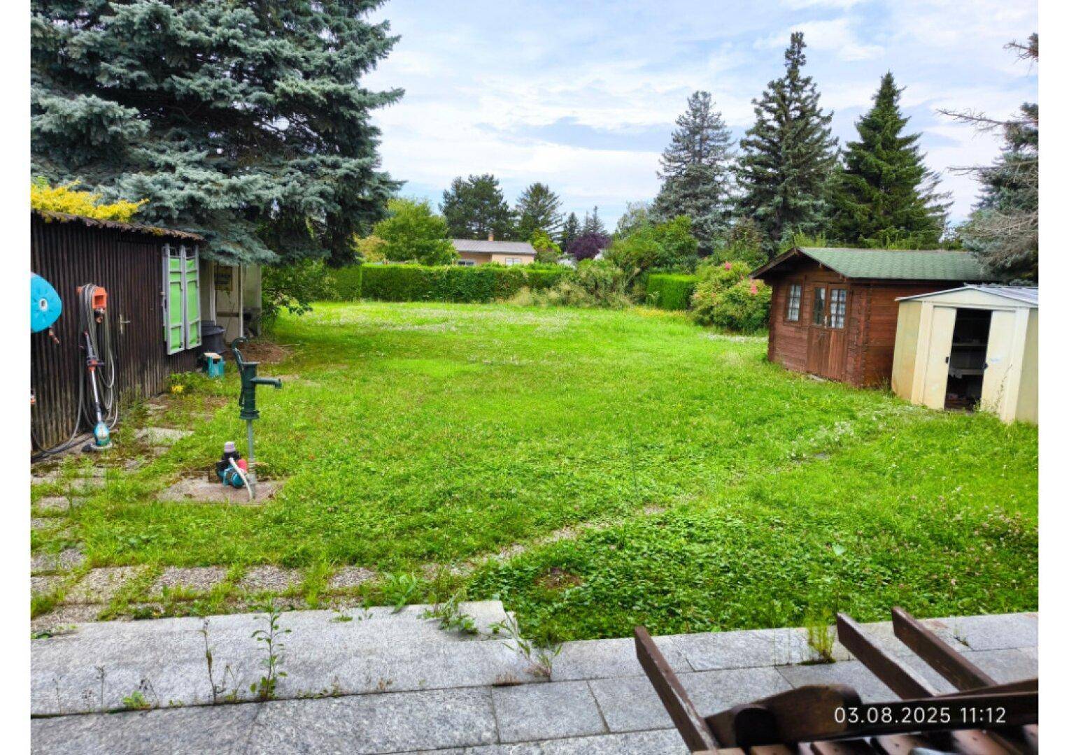 Großer Garten mit Rasen, Bäumen und Gartenhäusern, von einer Terrasse aus gesehen.
