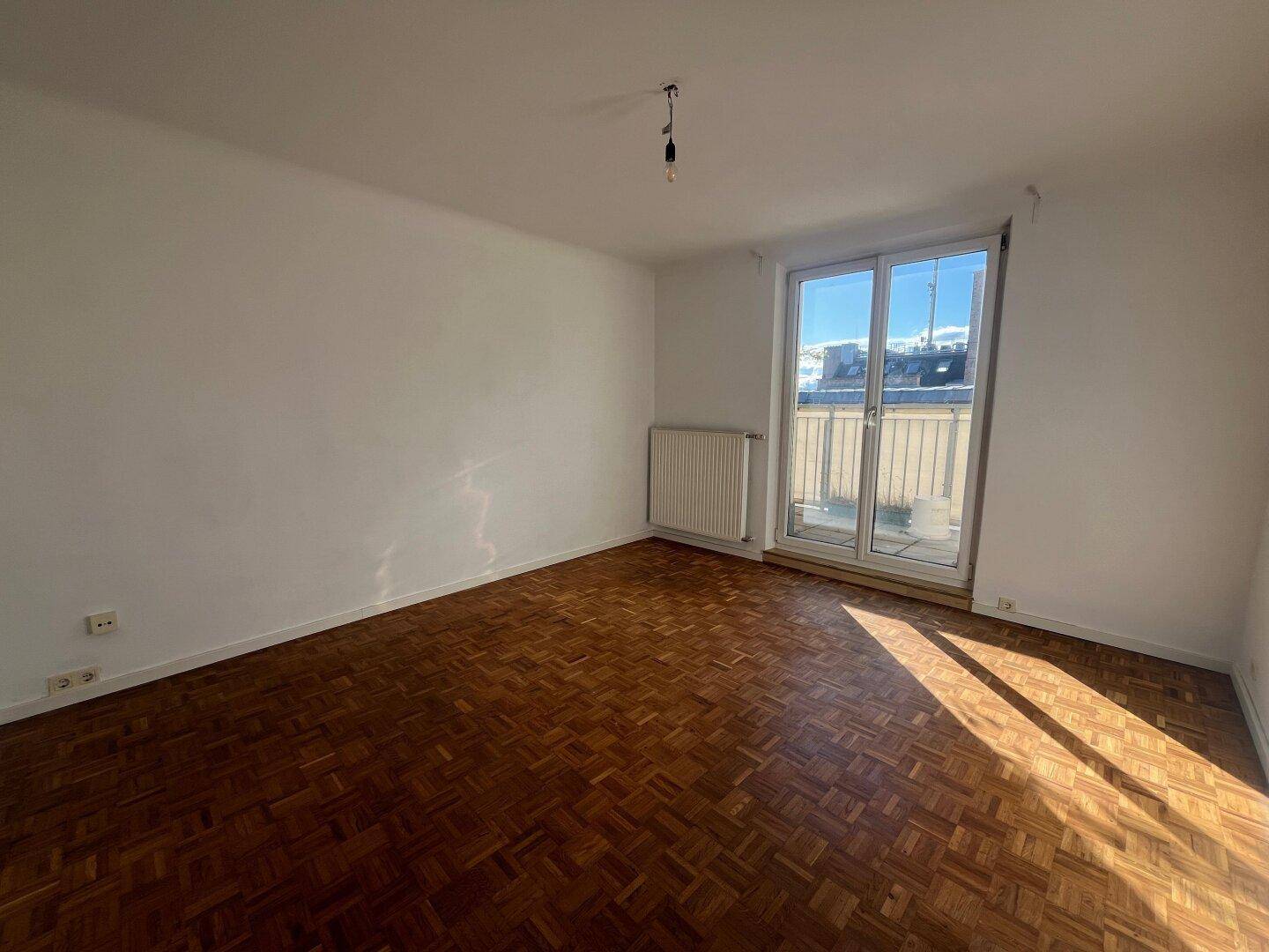 Leeres Wohnzimmer mit Fischgrätparkettboden und Zugang zu einem sonnigen Balkon.
