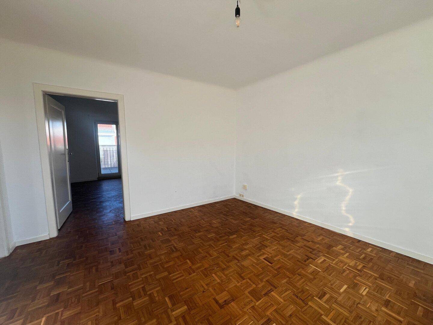 Helles Zimmer mit Parkettboden und Zugang zu einem angrenzenden Raum mit Balkon.