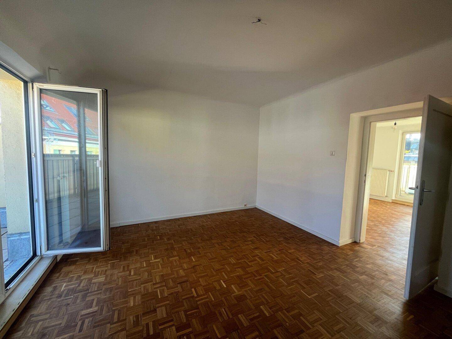 Helles Zimmer mit Parkettboden, Balkonzugang und offenem Durchgang zu einem weiteren Raum.
