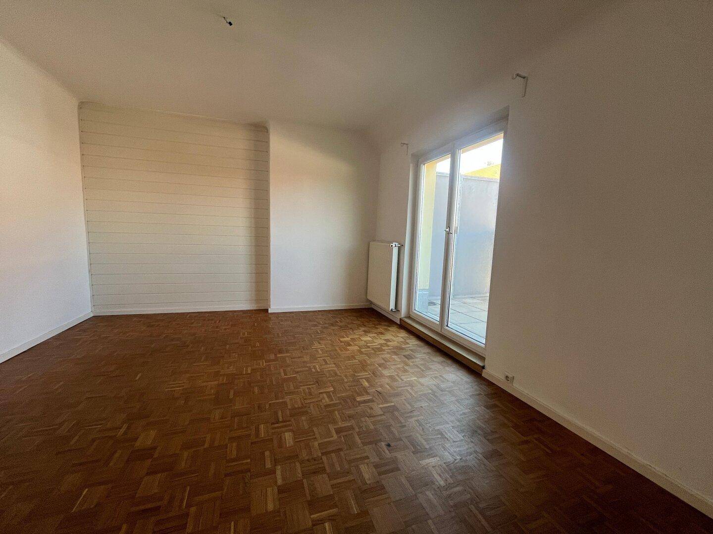 Geräumiges Zimmer mit Parkettboden und großen Fenstertüren, die auf einen Balkon führen.