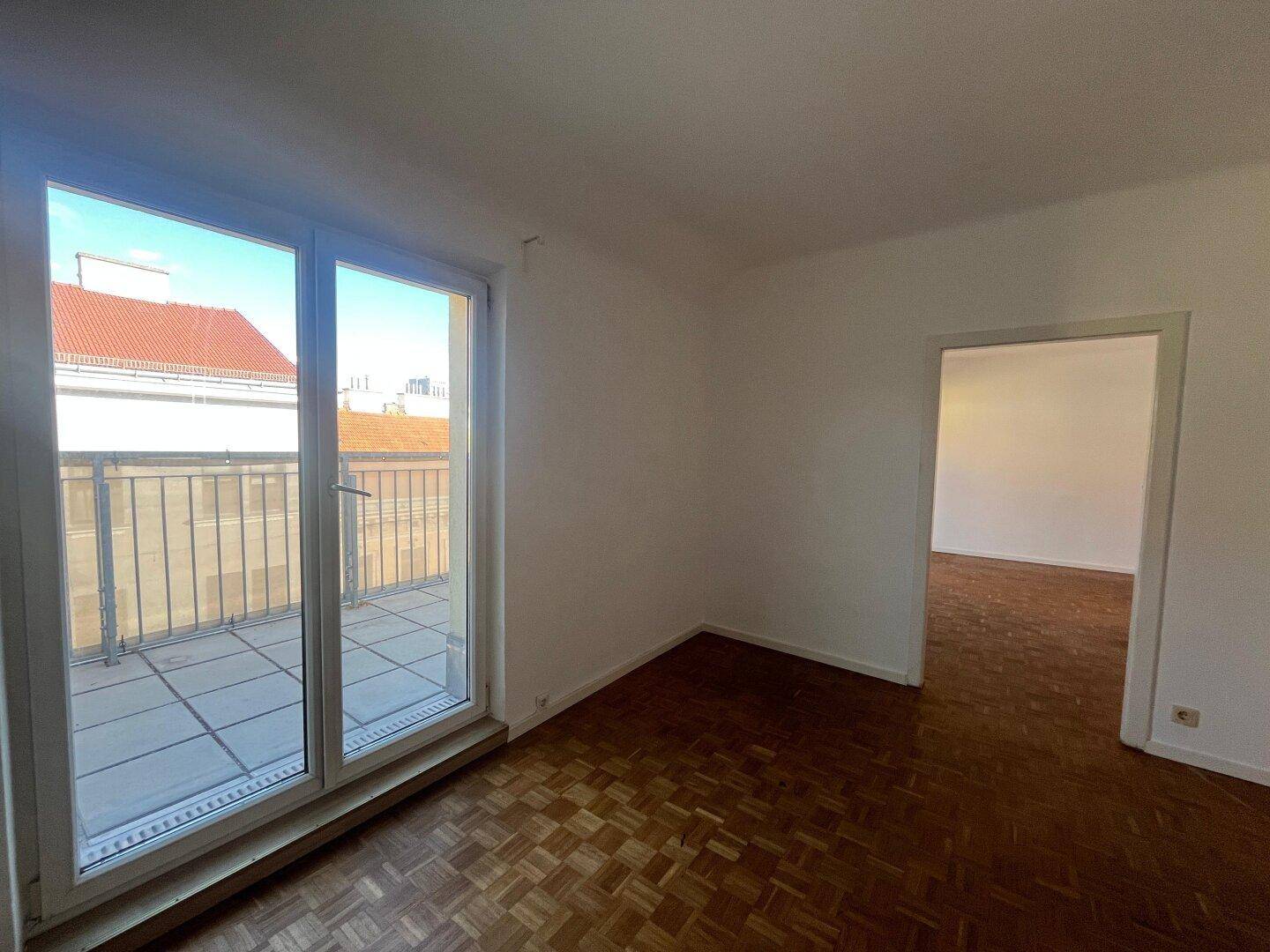 Helles Zimmer mit Parkettboden und großen Fenstern, die auf einen Balkon blicken.