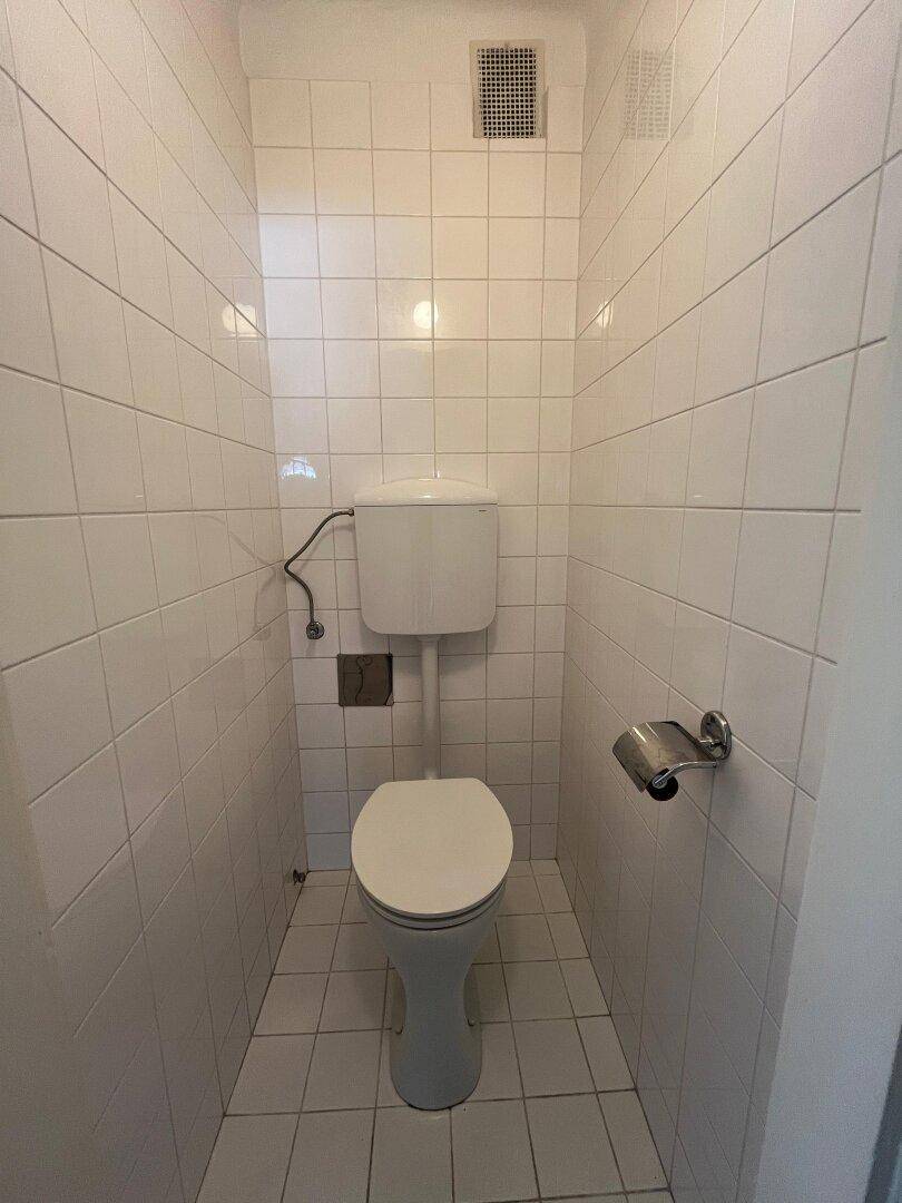 Kleines, vollständig gefliestes WC mit weißer Toilette und Lüftungsgitter.