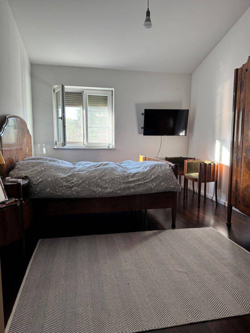 Gemütliches Schlafzimmer mit dunklem Holzfußboden, Fenster und Wandfernseher.