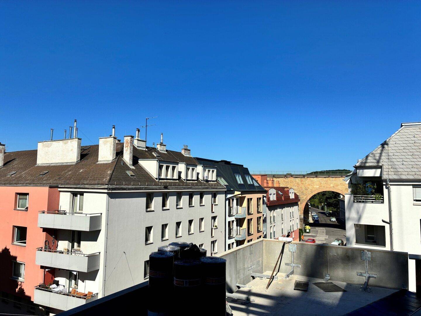 Blick über eine Wohnstraße mit Gebäuden und einer historischen Steinbrücke unter blauem Himmel.