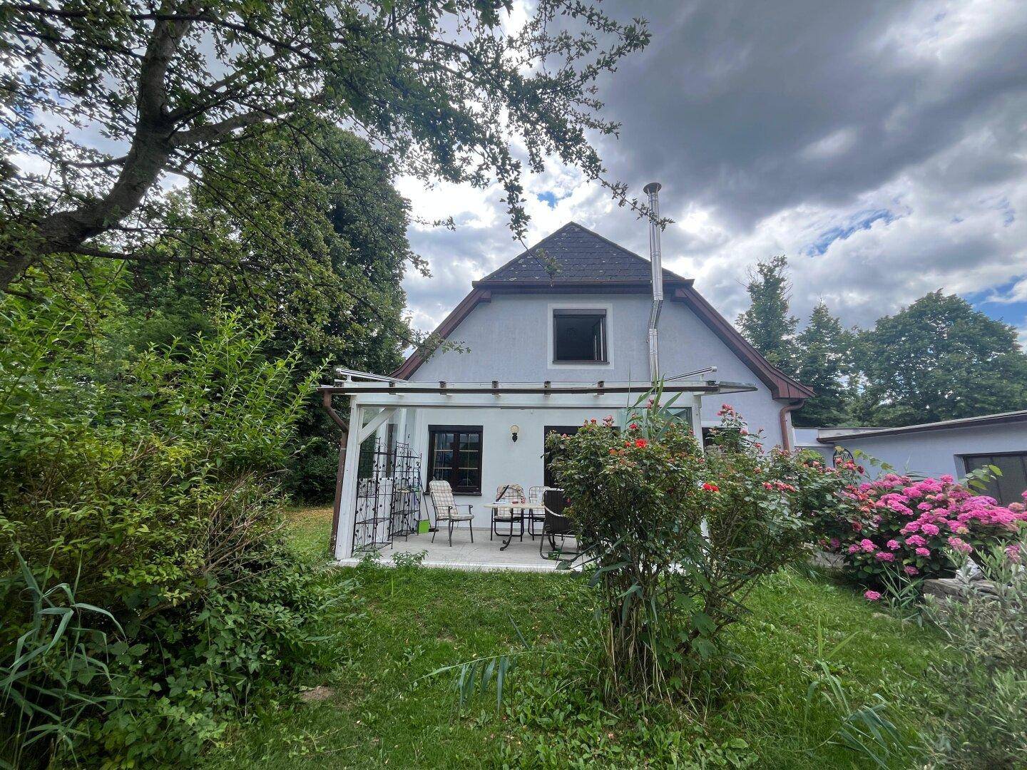 Haus mit überdachter Terrasse und blühenden Pflanzen im weitläufigen Garten.