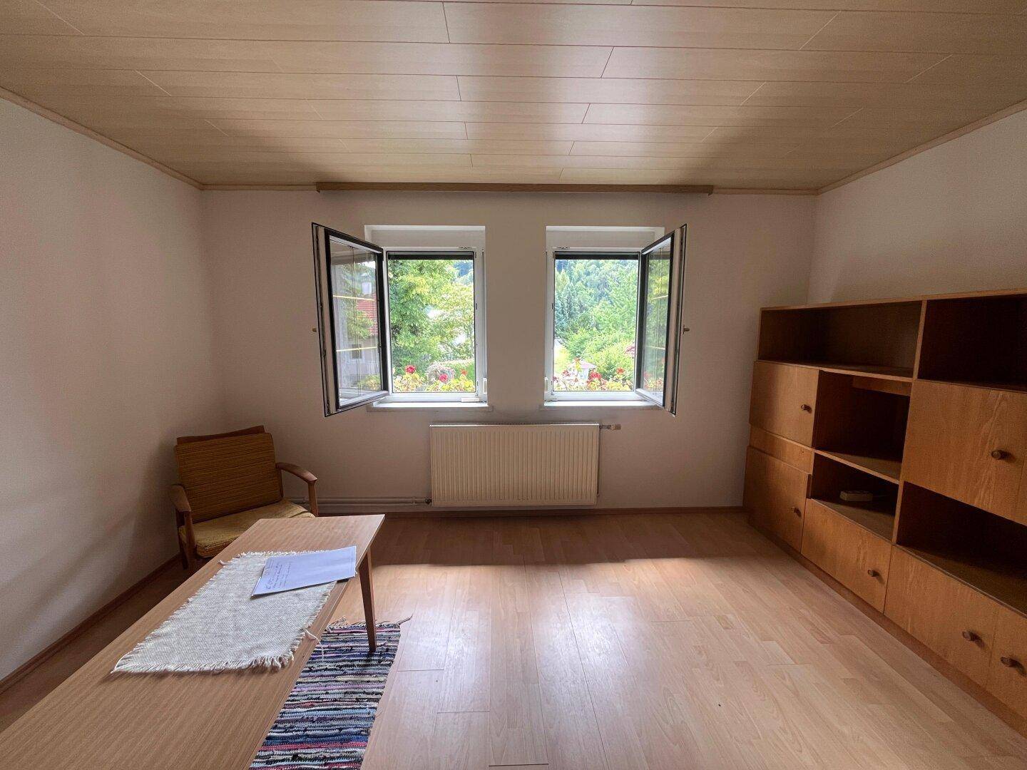 Helles Zimmer mit Holzboden, großen Fenstern und Blick auf die grüne Umgebung.