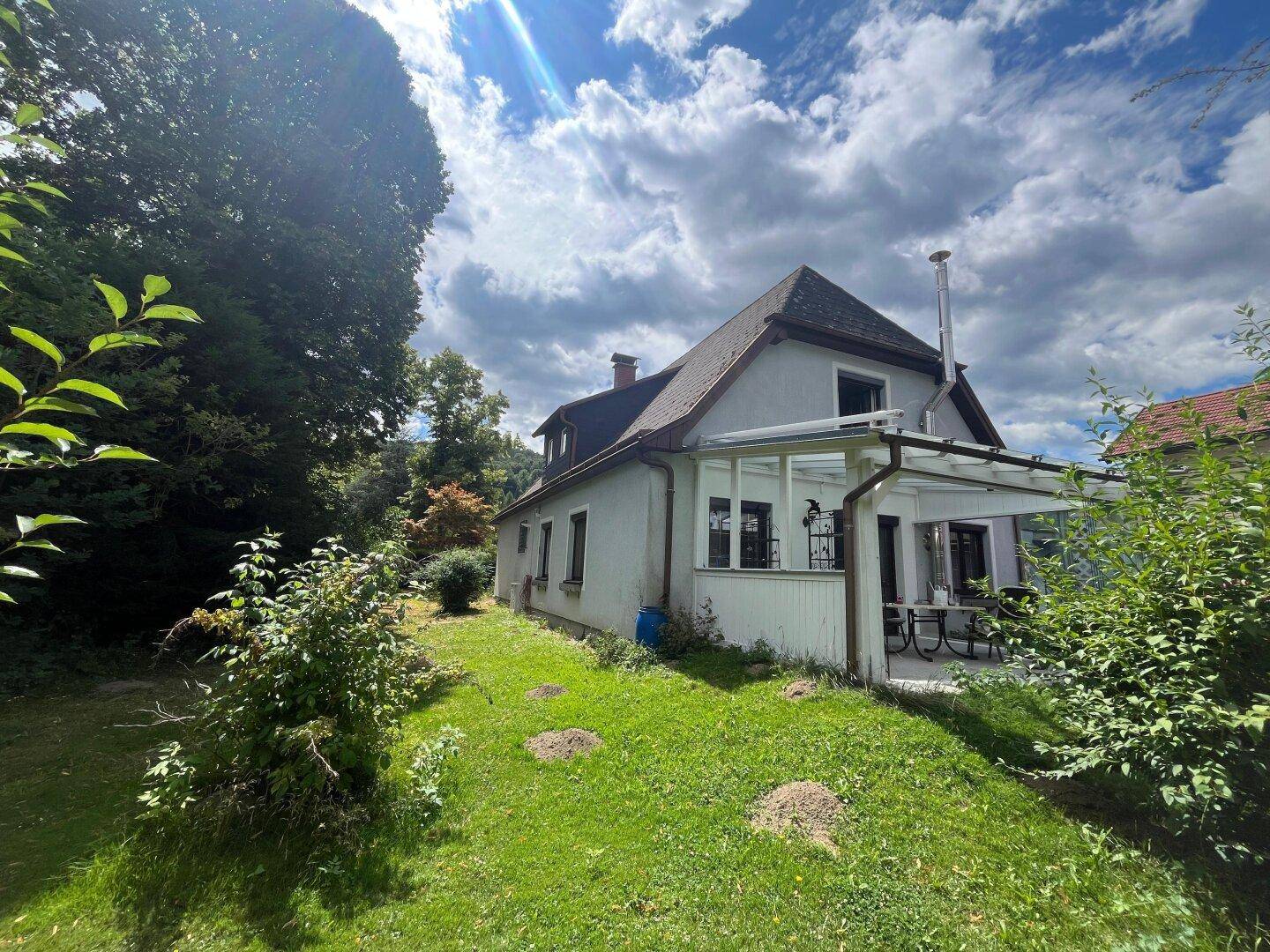 Hausfassade mit überdachter Terrasse und Gartenbereich unter bewölktem Himmel.