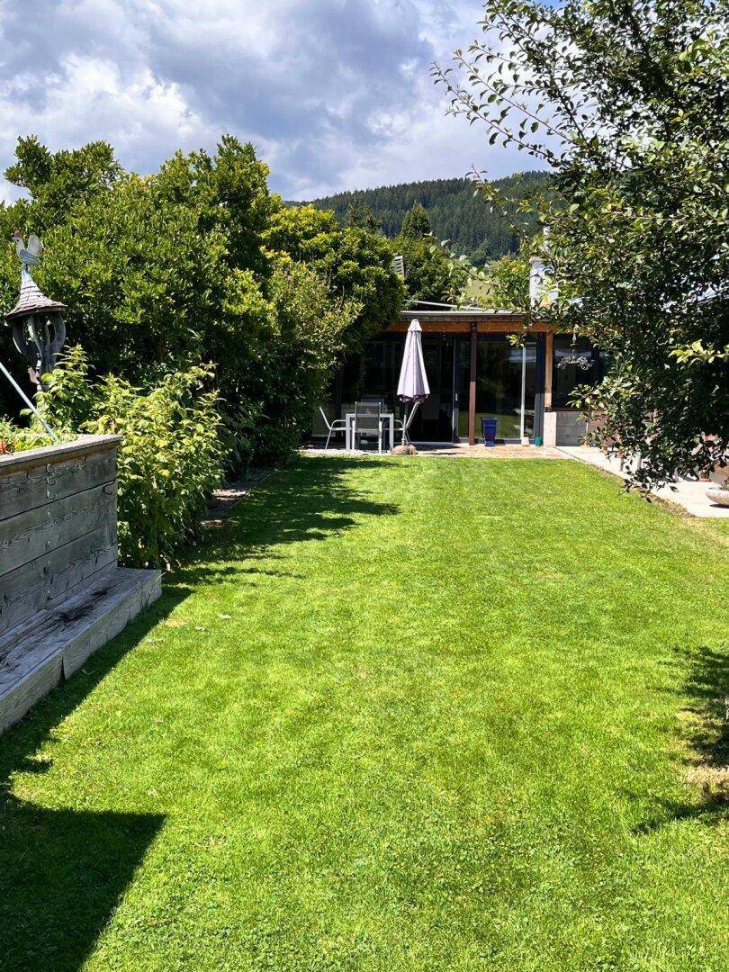 Grüner Gartenbereich mit Rasenfläche und einer möblierten Terrasse unter Bäumen.